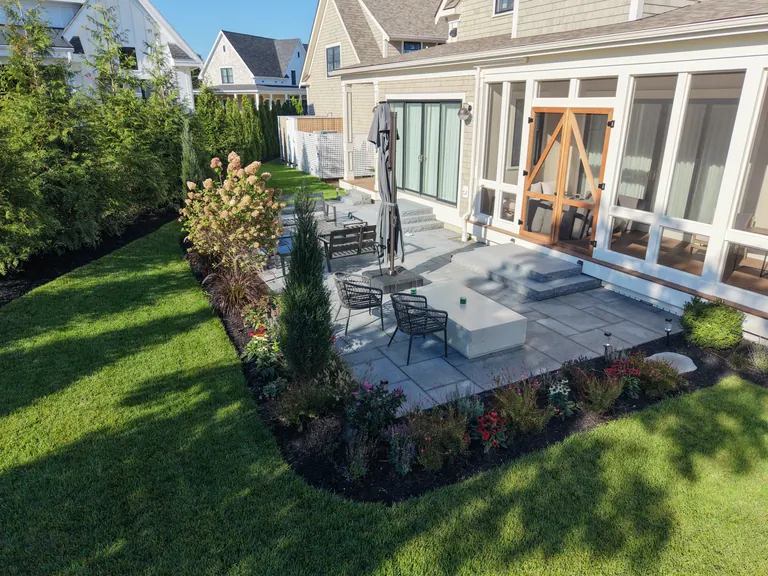 Furnished stone terrace with planting beside a gray-shingle home