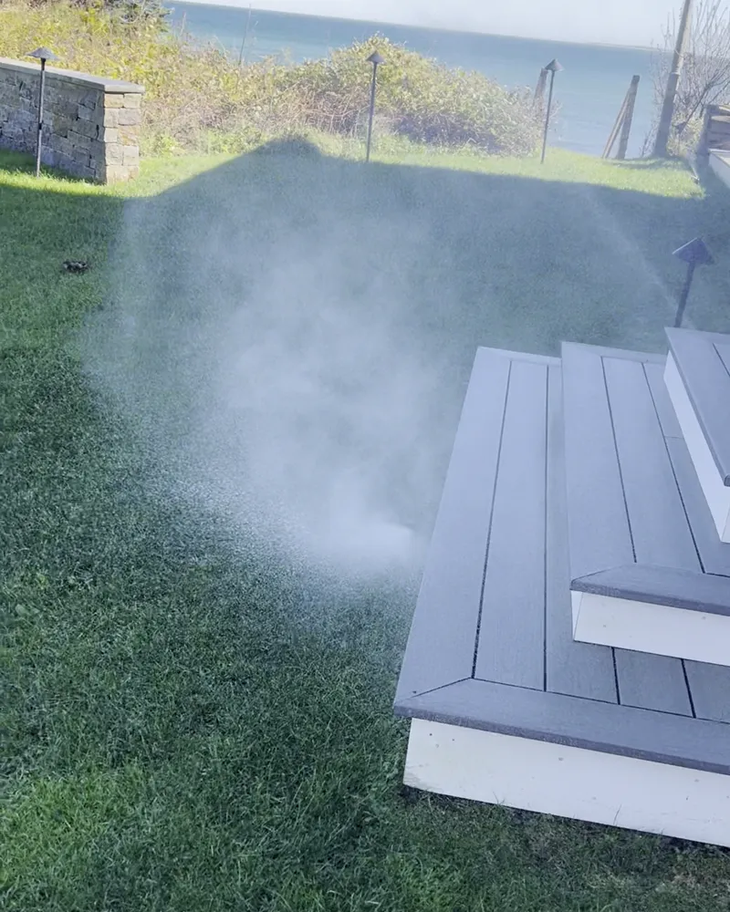Sprinkler heads misting on a waterfront lawn beside deck steps, with the ocean behind