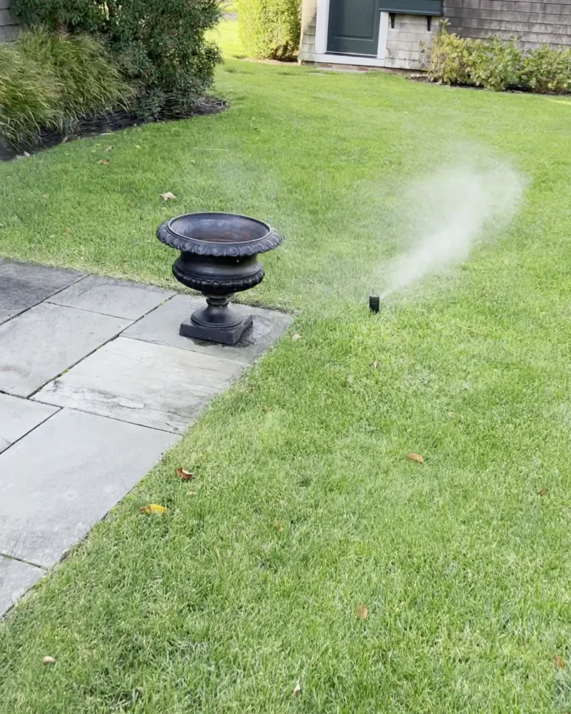 A sprinkler head misting as air clears the line, beside a stone patio and urn