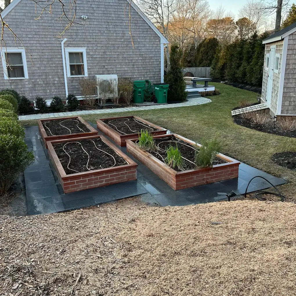Planted raised vegetable beds on a bluestone base behind a shingled house